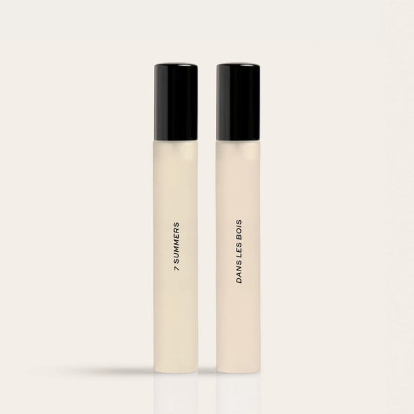 Two cylindrical fragrance bottles with black caps and cream-colored liquid, labeled "7 Summers" and "Damselless Bodies".