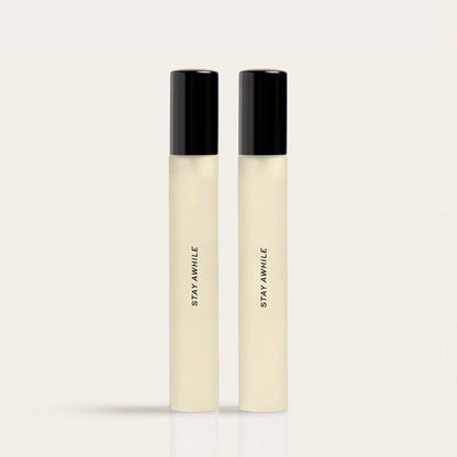 Two cylindrical fragrance bottles with black caps and pale liquid, labeled "STAY AWHILE."